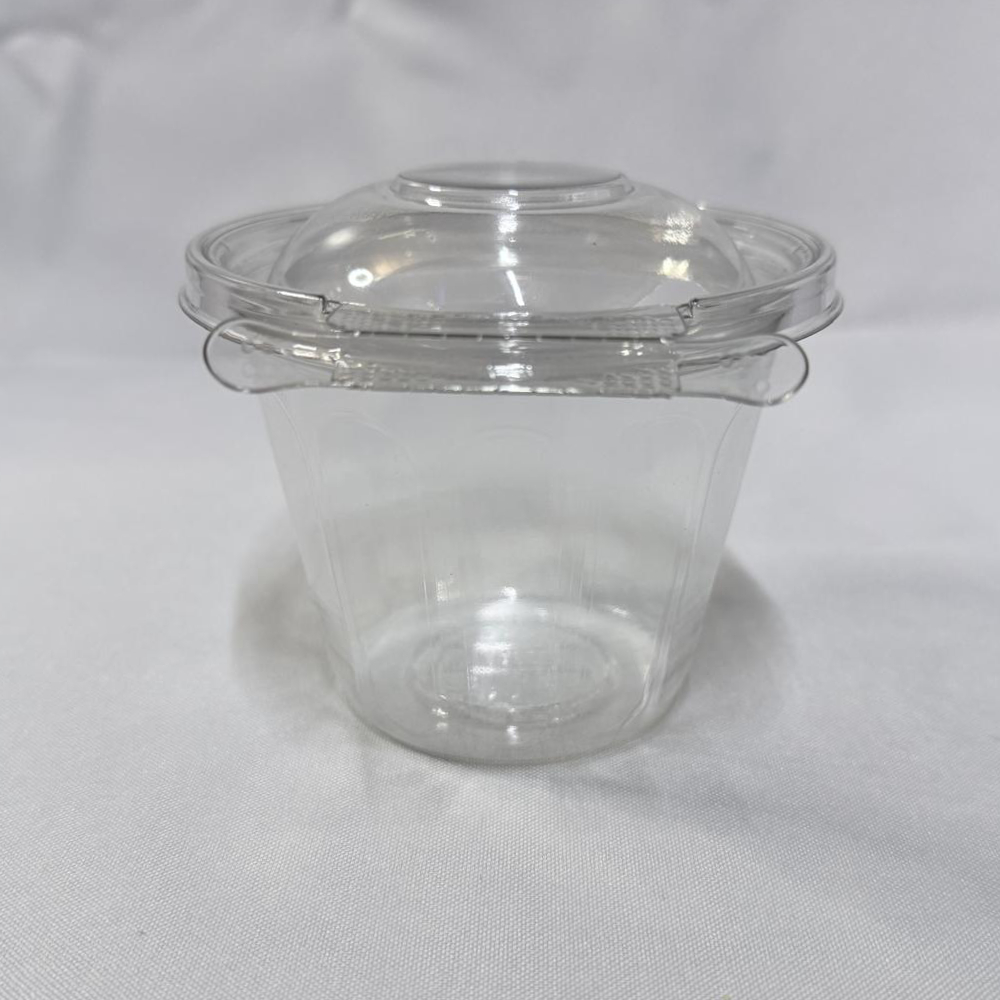 Envase redondo descartable de plástico transparente (PET) tipo estuche con base profunda y tapa chata. Se presenta en dos imágenes: una con el envase completamente abierto mostrando el interior de su cavidad profunda y la tapa unida por bisagra en segundo plano, y otra del envase cerrado visto de frente donde destaca la pestaña del precinto de seguridad perimetral, ambas sobre un fondo claro.