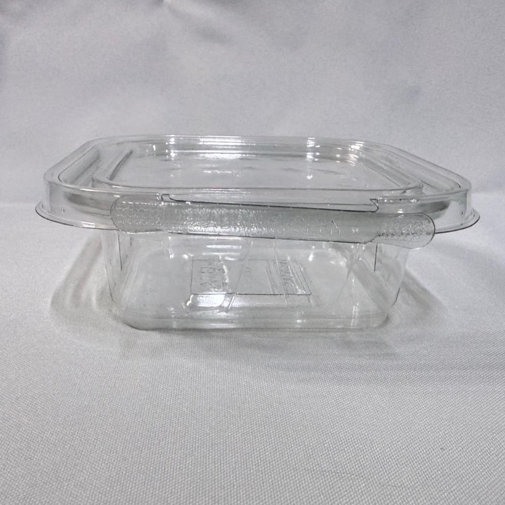 Envase cuadrado descartable de plástico transparente (PET) tipo estuche. Se presenta en dos imágenes: una mostrando el envase completamente abierto y visto en ángulo desde arriba para apreciar su interior profundo y la tapa unida por bisagra, y otra del envase cerrado visto de frente donde resalta la pestaña del precinto de seguridad con la inscripción "ABRA / OPEN", ambas sobre un fondo claro.