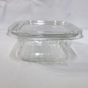 Envase cuadrado descartable de plástico transparente (PET) tipo estuche. Se presenta en dos imágenes: una mostrando el envase completamente abierto y visto en ángulo desde arriba para apreciar su interior y la tapa unida por bisagra, y otra del envase cerrado visto de frente donde resalta la pestaña frontal del precinto de seguridad, ambas sobre un fondo claro.
