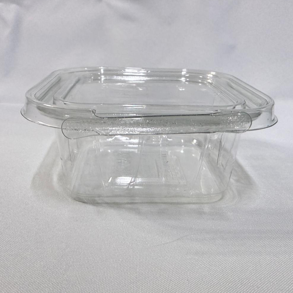 Envase cuadrado descartable de plástico transparente (PET) tipo estuche. Se presenta en dos imágenes: una mostrando el envase completamente abierto y visto en ángulo desde arriba para apreciar su interior y la tapa unida por bisagra, y otra del envase cerrado visto de frente donde resalta la pestaña frontal del precinto de seguridad, ambas sobre un fondo claro.
