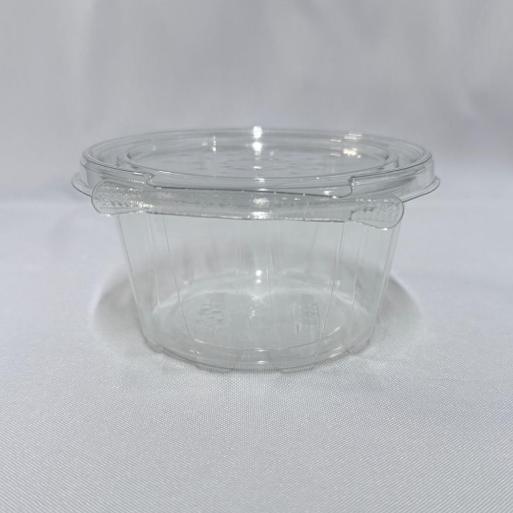 Envase redondo descartable de plástico transparente (PET) tipo estuche. Se presenta en dos imágenes: una mostrando el envase cerrado en vista frontal donde se destaca la pestaña del precinto de seguridad, y otra con el envase abierto mostrando su tapa unida por bisagra en segundo plano, ambas sobre un fondo claro.