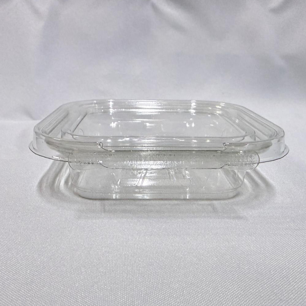 Envase rectangular descartable de plástico transparente (PET) tipo estuche. Se presenta en dos imágenes: una mostrando el envase completamente abierto con su tapa unida por bisagra, y otra cerrado en vista frontal donde se destaca la pestaña del precinto de seguridad, ambas sobre un fondo claro.