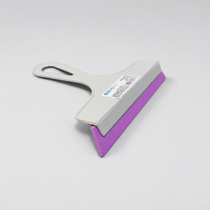 Vista superior plana de un mini lampazo o secador de mano con mango de plástico gris y labio de goma color fucsia, mostrando la etiqueta del fabricante adherida en el dorso, sobre un fondo gris claro.