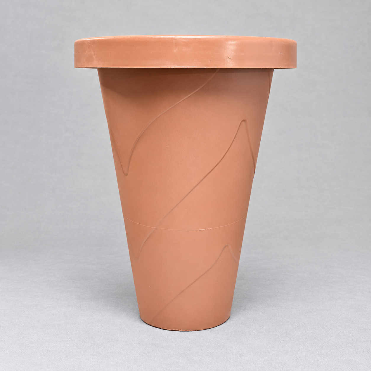 Vista frontal de una maceta de plástico alta y de forma cónica en color terracota, destacando un borde superior grueso y líneas curvas en relieve que decoran su superficie, fotografiada sobre un fondo gris claro.